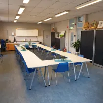 Een creatieve en multifunctionele ruimte met een open en lichte sfeer. De lange tafels in een U-vorm opstelling bieden plaats aan groepsactiviteiten zoals workshops, vergaderingen of cursussen. De blauwe stoelen geven een kleurrijke touch aan de ruimte. Aan de achterkant bevindt zich een kitchenette, handig voor het bereiden van koffie of thee. De kunstwerken en schildersezels langs de muur wijzen op een creatieve omgeving, ideaal voor kunst- en handvaardigheidssessies.
