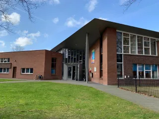 Een modern gebouw met een bakstenen gevel en grote glaspartijen bij de entree, wat zorgt voor een open en uitnodigende uitstraling. Aan de voorzijde loopt een strak betegeld pad langs een groene grasstrook en een grote boom die voor schaduw zorgt. Posters en borden naast de ingang duiden op activiteiten of informatie binnen het gebouw. De combinatie van natuur en architectuur creëert een rustige, aangename sfeer.